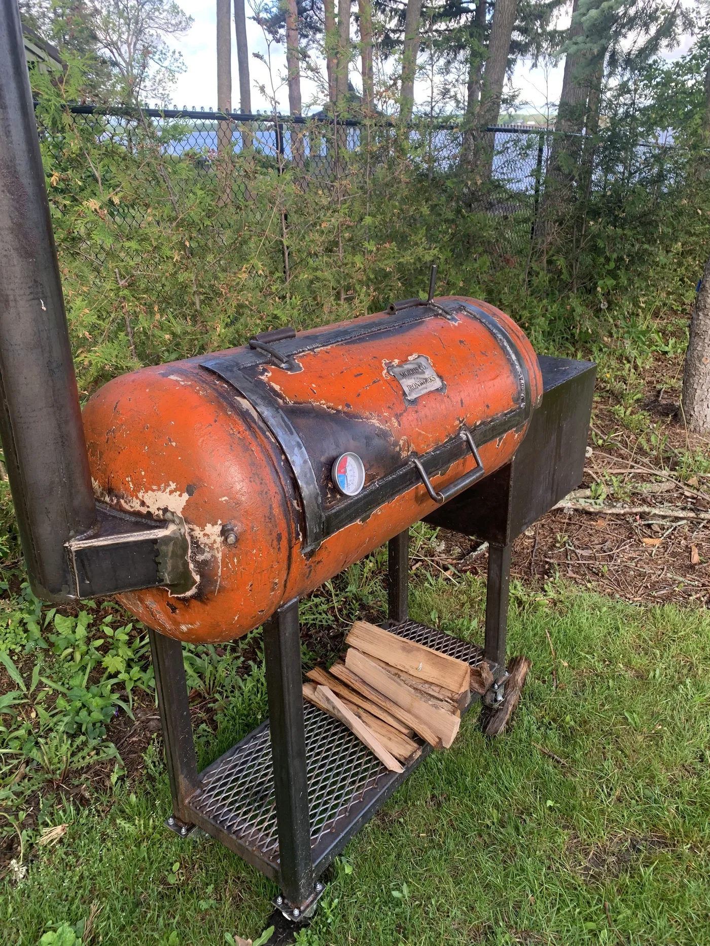 The orange Morris Co. Ironworks offset smoker on the lawn, with oak splits stacked on the rack underneath