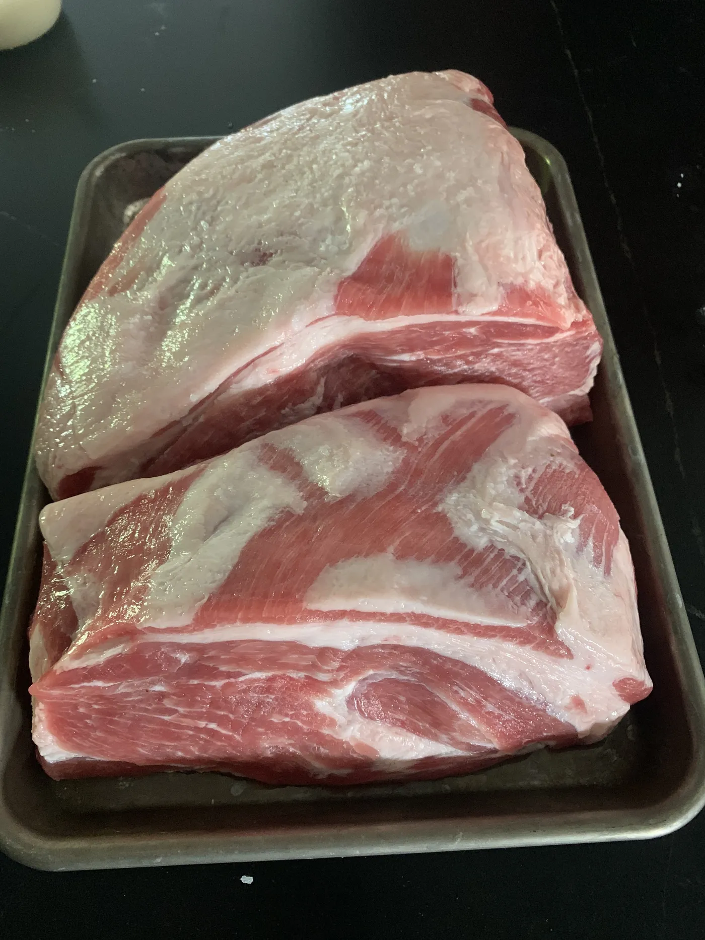 Two raw halves of pork butt, fat side up, on a metal sheet pan