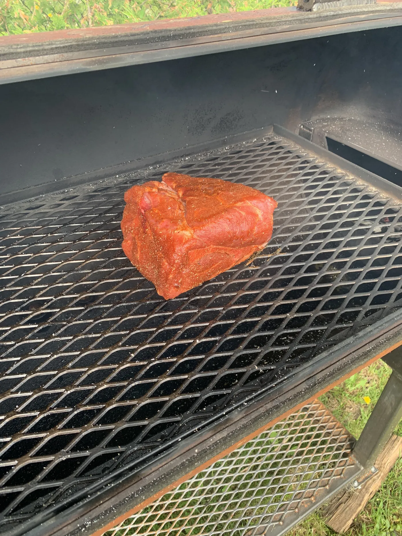 A piece of pork butt coated in red Memphis Dust rub on the grate of the offset smoker's cook chamber